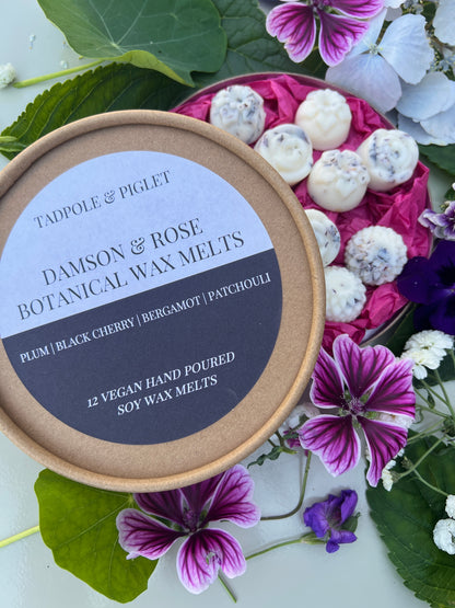 Damson &amp; Rose Botanical Wax Melts in a round container surrounded by flowers and greenery, showcasing a luxurious fragrance.