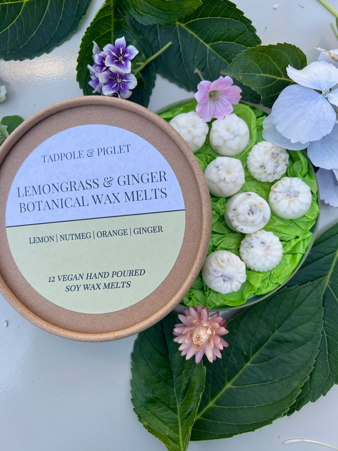 Lemongrass &amp; Ginger Botanical Wax Melts in a brown container, surrounded by fresh flowers and green leaves. Vegan hand poured soy wax melts.