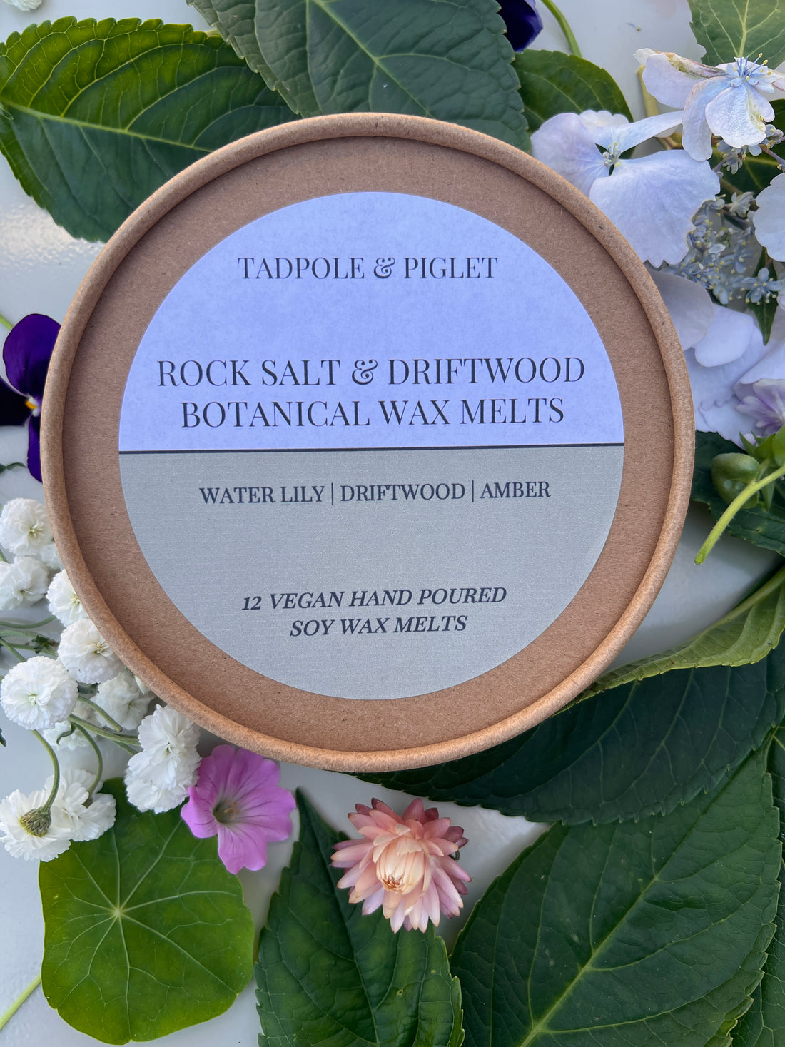 Rock Salt &amp; Driftwood Botanical Wax Melts in a round cardboard container surrounded by flowers and leaves.