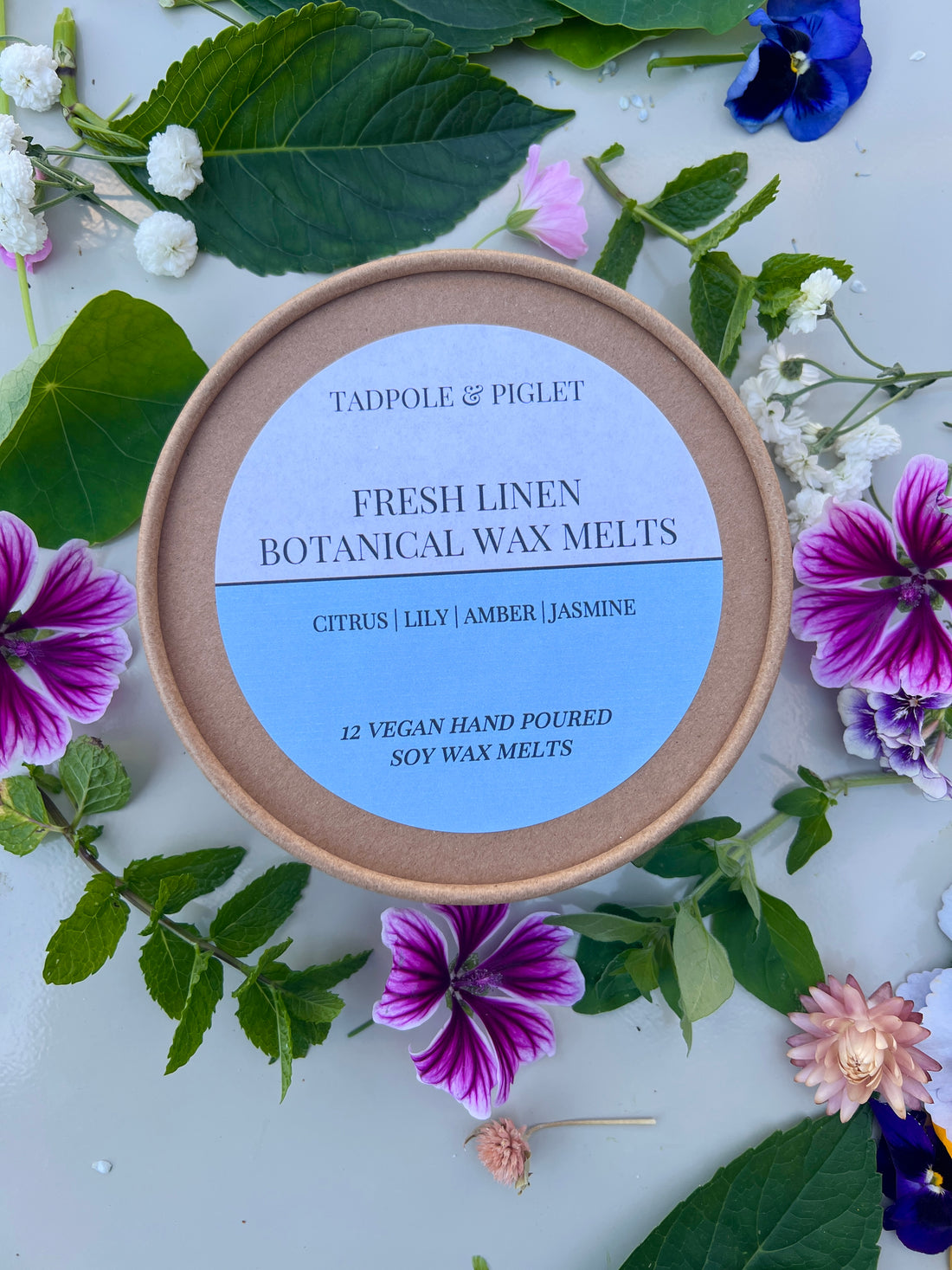 Fresh Linen Botanical Wax Melts in round packaging surrounded by fresh flowers and leaves.