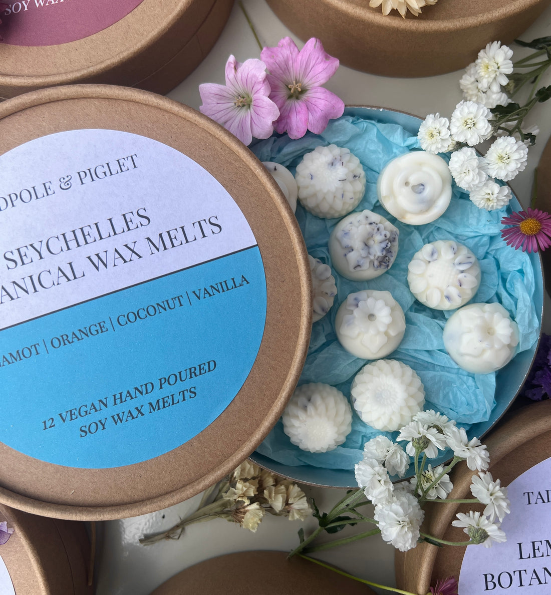 Seychelles Botanical Wax Melts in a round box surrounded by flowers, featuring bergamot, orange, coconut, and vanilla fragrances.