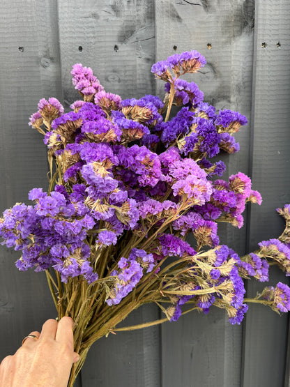 Bunch of vibrant purple statice flowers held against a wooden background, showcasing their rich color and traditional charm.